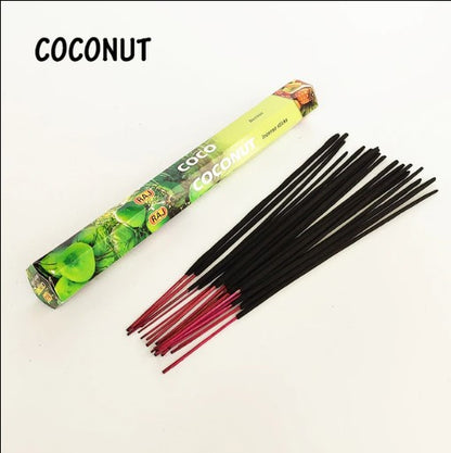 A bundle of dark incense sticks with red tips laid out next to a green and yellow rectangular box labeled "Coconut" with images of coconut on it, on a plain white surface.